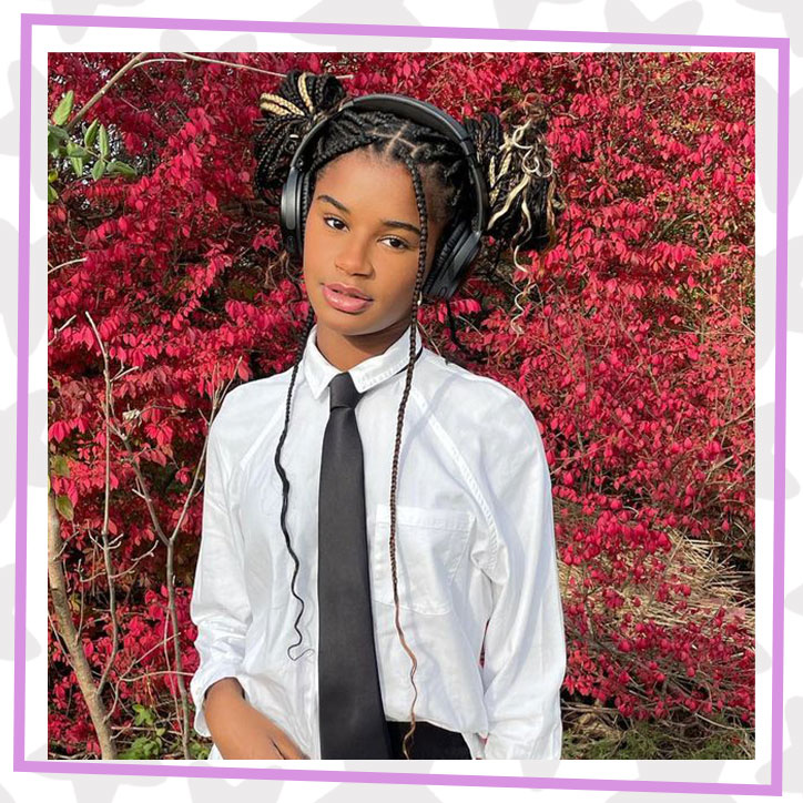 Marley Dias standing in front of a tree with red leaves. She is wearing a white button down shirt and long black tie. She has headphones on and her braided hair is up in messy buns.