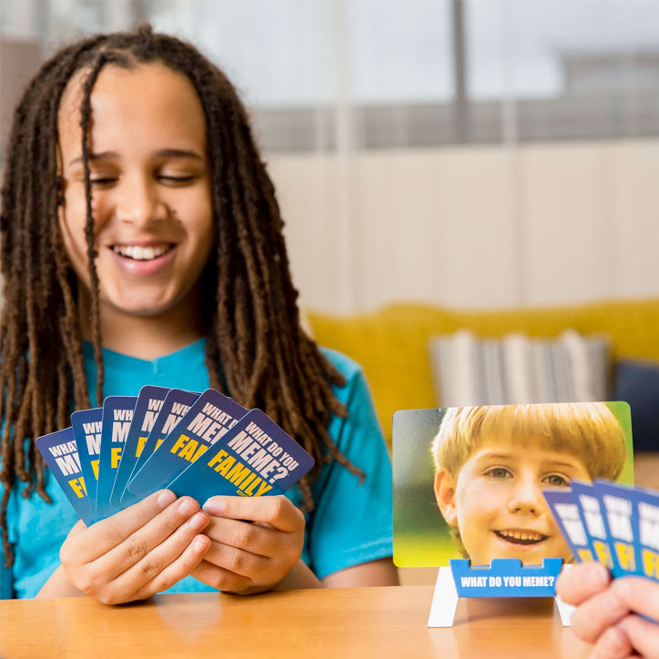 Lifestyle photo of kids playing What Do You Meme? Family Edition at a table. They are smiling and holding game cards.