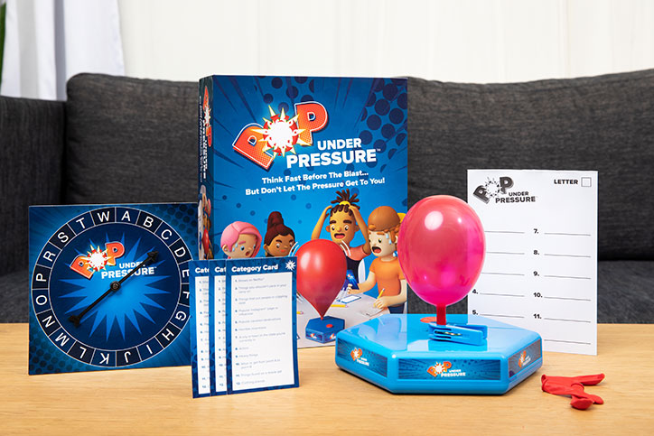 Lifestyle photo of the Pop Under Pressure game sitting on a living room table. Photo features the game box, balloon timer, category sheets, spinner, and other game elements