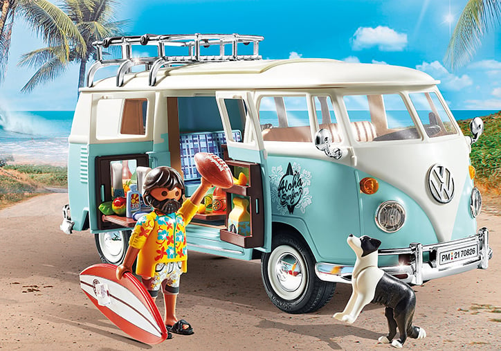 Lifestyle photo of an assembled Playmobil Volkswagen T1 Camping Bus Special Edition sitting on a model beach while the included figure holds the surfboard accessory and looks like he's going to throw the football accessory to the Border Collie figure