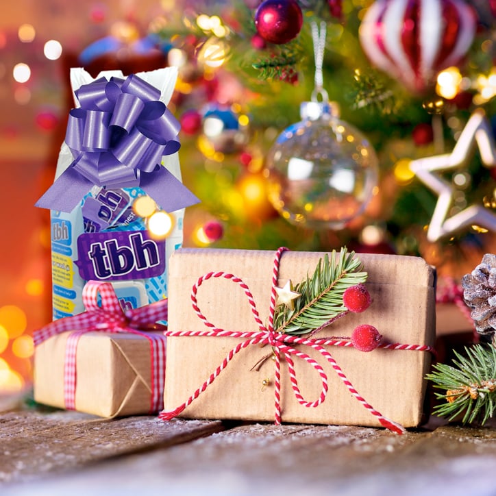 Picture of a TBH Kids Holiday Gift Set with a big purple bow nestled under a Christmas tree alongside other gifts