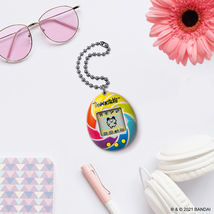 Flat lay of an Original Tamagotchi device laying on a table with aesthetic items like a pink pen, pink flower, pink sunglasses, and white headphones