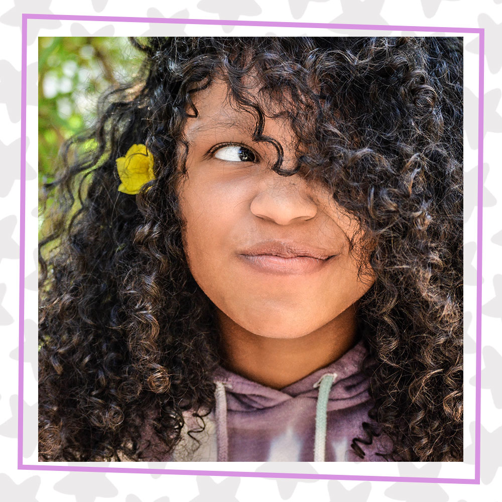 Closeup of Nadia Simms with a scrunched nose, looking up to the left, her curly hair falling in front of her face. She is wearing a tye dye hoodie and a yellow flower in her hair.