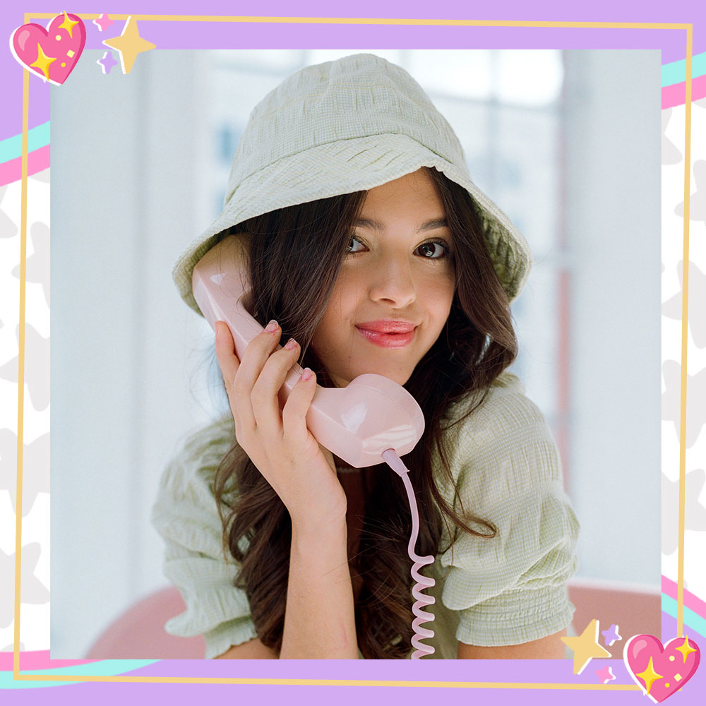 Adriana Camposano poses in a blush pink chair, casually holding a retro telephone. She is wearing a mint green dress and bucket hat