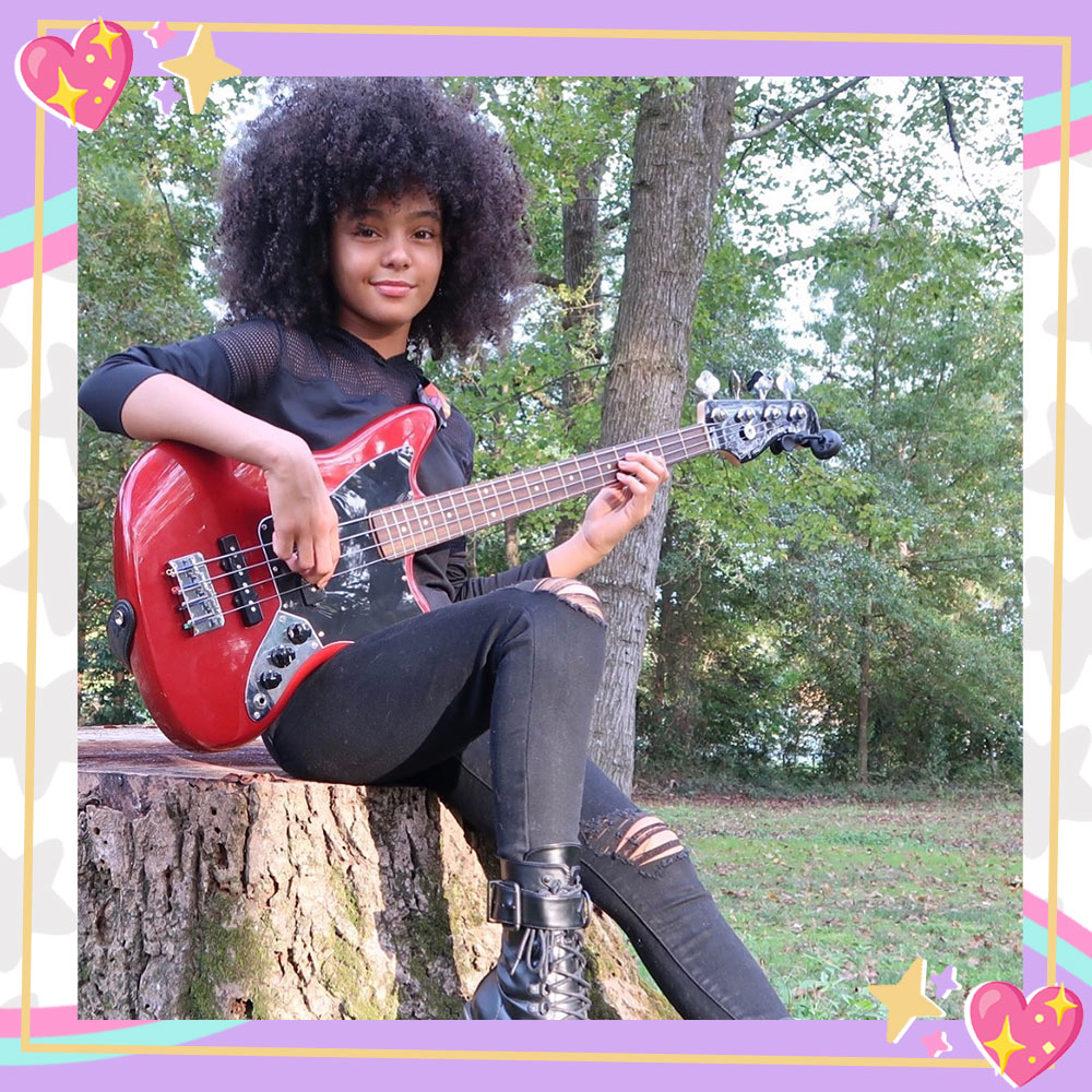 Madalen Mills sits on a tree stump in a wooded area. She is holding a red electric guitar. She is wearing her natural curls, ripped black jeans, black combat boots, and a black fishnet top.