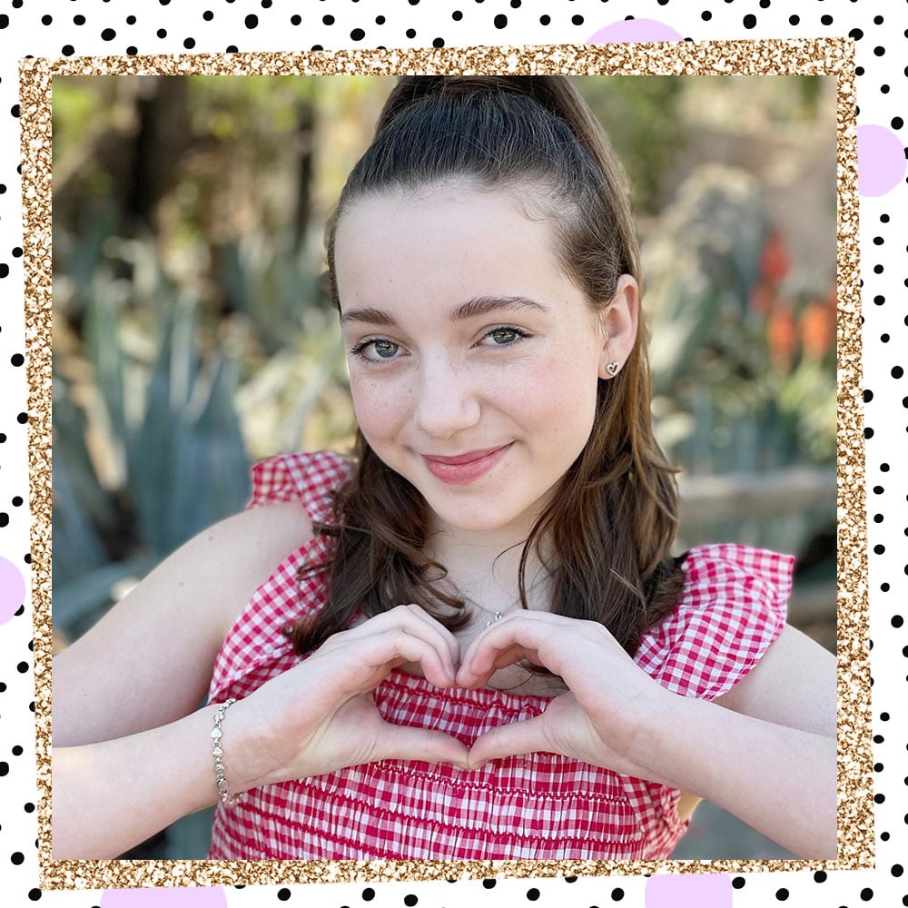 Photo of Libby Rue outside. She is wearing a red and white gingham top with ruffled short sleeves, making a heart with her hands