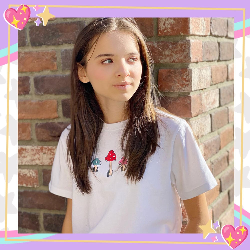 Madeleine McGraw poses in front of a brick wall, looking off to the side. She is wearing a white t-shirt with illustrated mushrooms on it.
