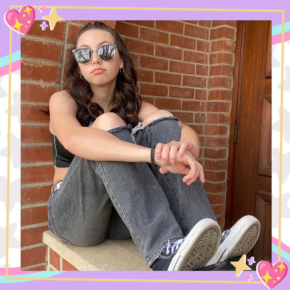 Lola Raie sits up on a ledge in front of a brick wall. She has her knees pulled up to her chest with her arms wrapped around them. She is wearing big sunglasses, ripped black jeans, and converse sneakers.