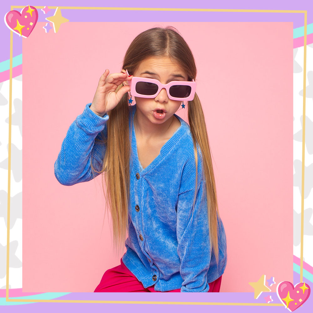 Mandy Corrente poses in front of a pink backdrop. She is wearing a blue crushed velvet cardian, baby pink oversized sunglasses, and her hair is in pigtails.