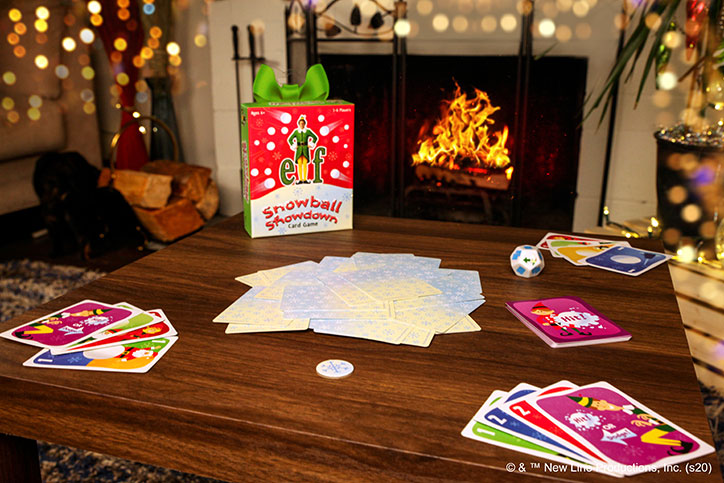 Lifestyle photo of the Elf: Snowball Showdown card game set up around a living room table. There are holiday lights and a fire in the background.