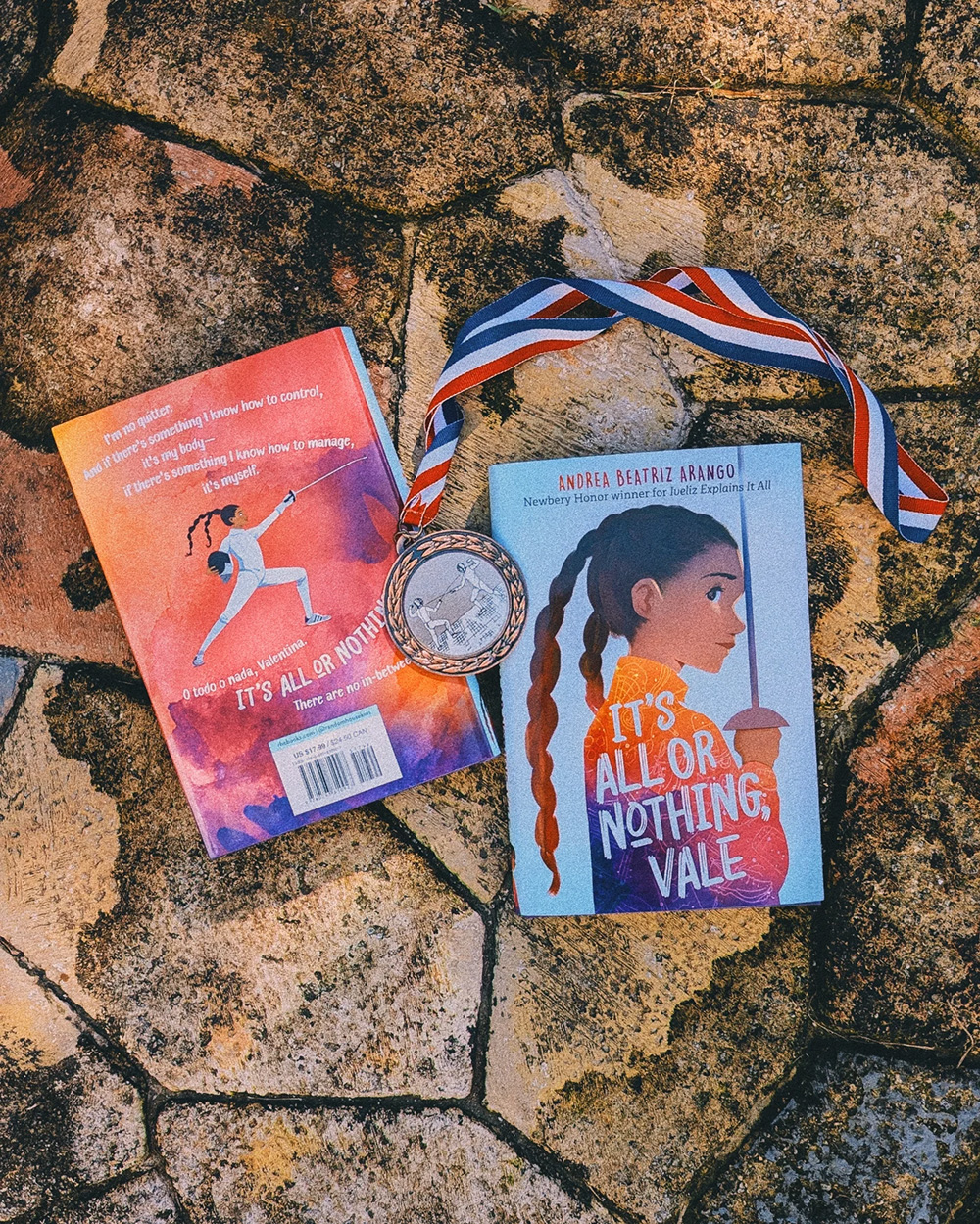 Two hardcover copies of It's All or Nothing, Vale on the ground, showcasing both the front and back covers. There is a gold medal for fencing laid across them.