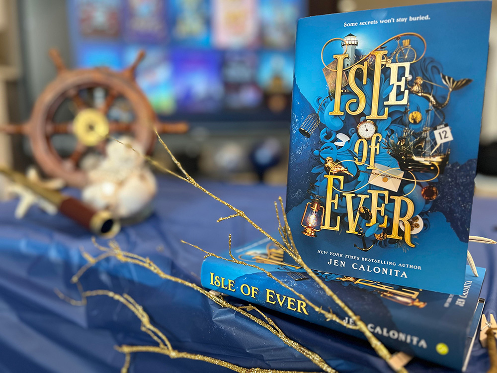 A stack of hardcover copies of Isle of Ever on their sides, with one book standing on top. There are golden branches, a pirate ship helm, spyglass, coral, and a starfish arranged decoratively around the books.