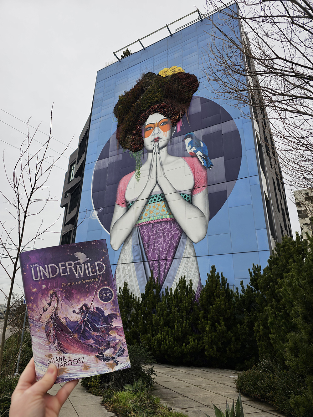 Photo of the Attitude of Gratitude mural in Portland, Oregon. Someone is holding up a copy of The Underwild: River of Spirits in front of the mural.