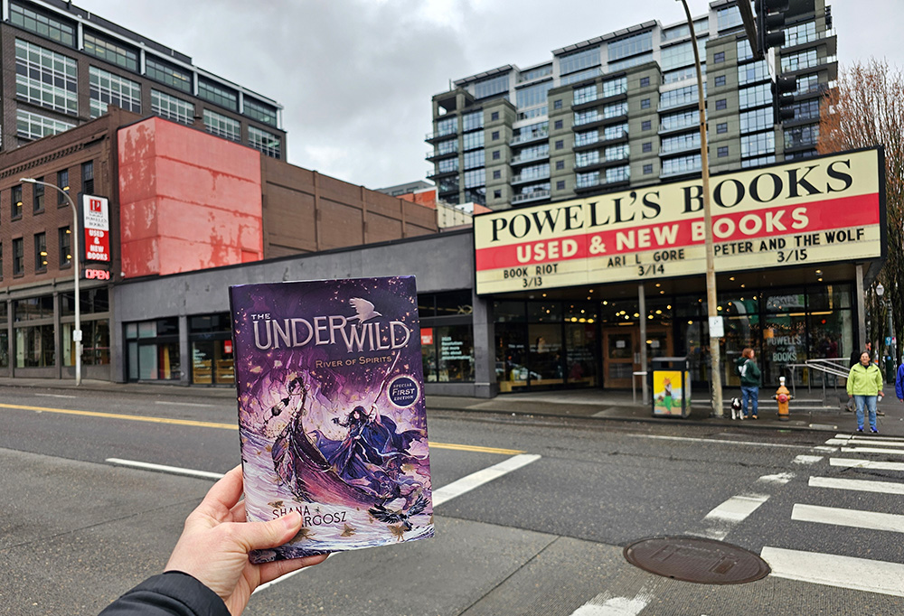 Photo of the iconic Powell's Books bookstore in Portland, Oregon. A hand is holding up a copy of The Underwild: River of Spirits in the shot.